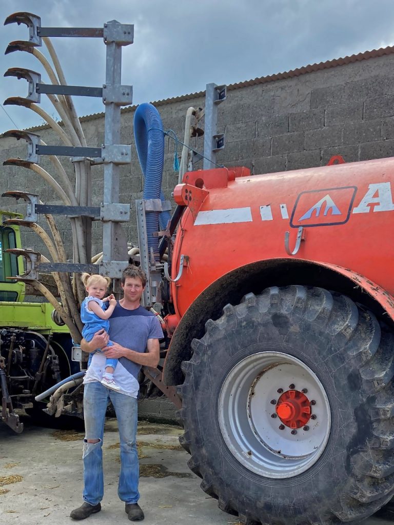 Father holding toddler in arms beside red slurry tanker
